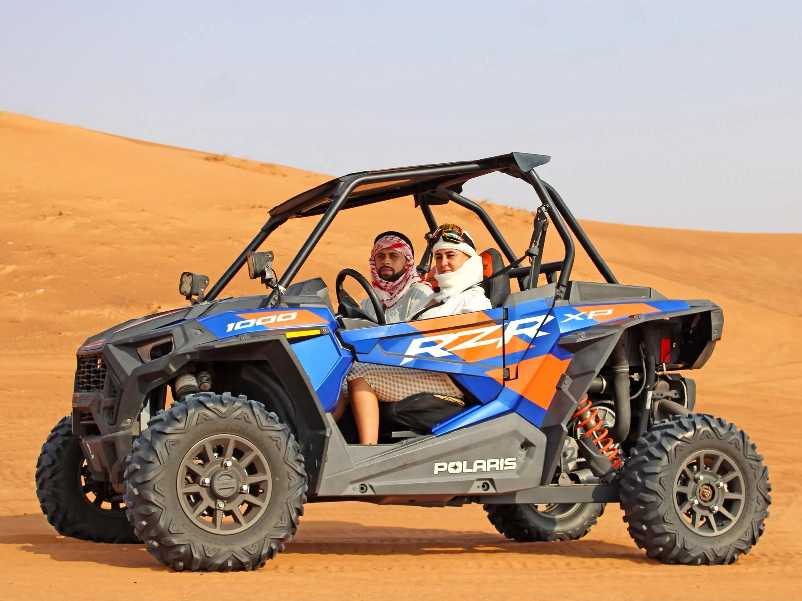Couple driving a blue Polaris RZR 1000 dune buggy through the Dubai desert